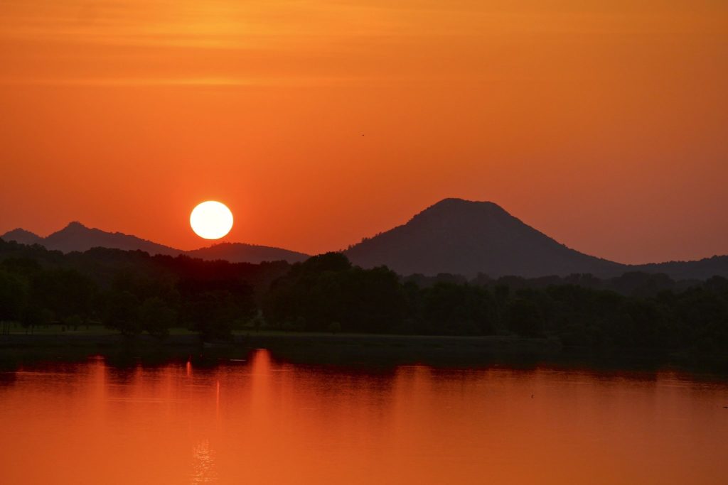 Image shows the sun setting over Pinnacle Mountain. The sky is orange, and the water is reflecting the orange color.