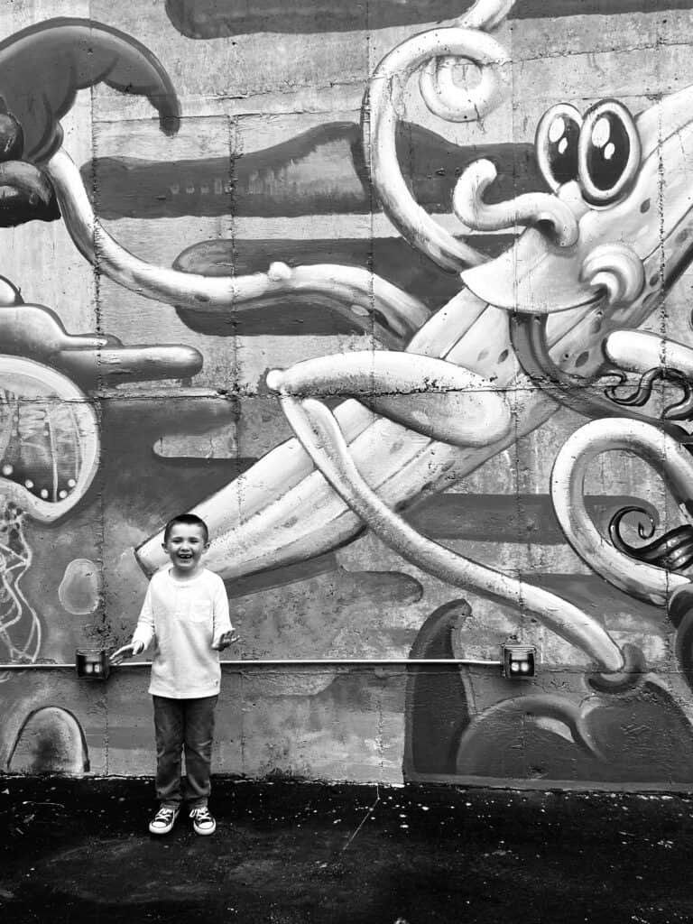 Black and white image of a boy smiling in front of a large mural of a silly banana