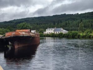 rusty old boat sits in water. a house is on the shore in the background
