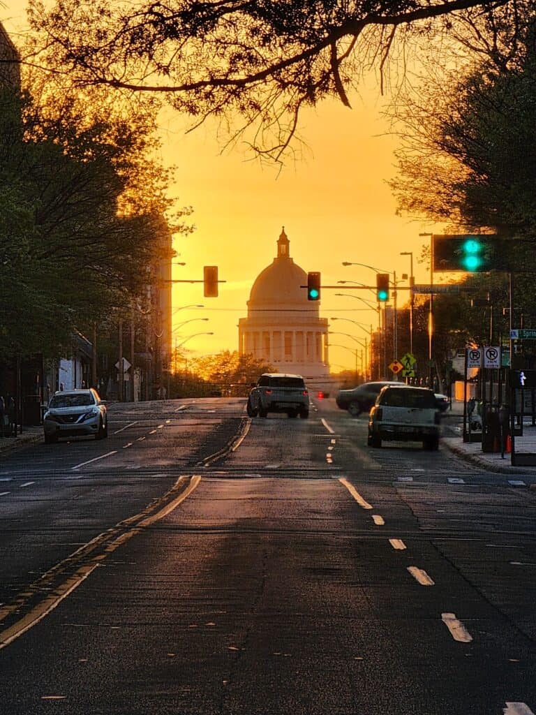 The Arkansas State Capitol dome is seen during golden hour. Vehicles on Capitol Avenue, which leads to the building, are seen on the street in the foreground.