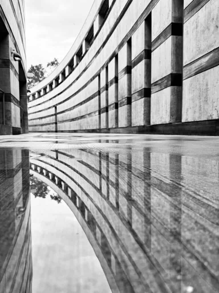 black and white, a curved brutalist or modernist building is reflected in water on the ground. The camera angle is low, close to the ground.