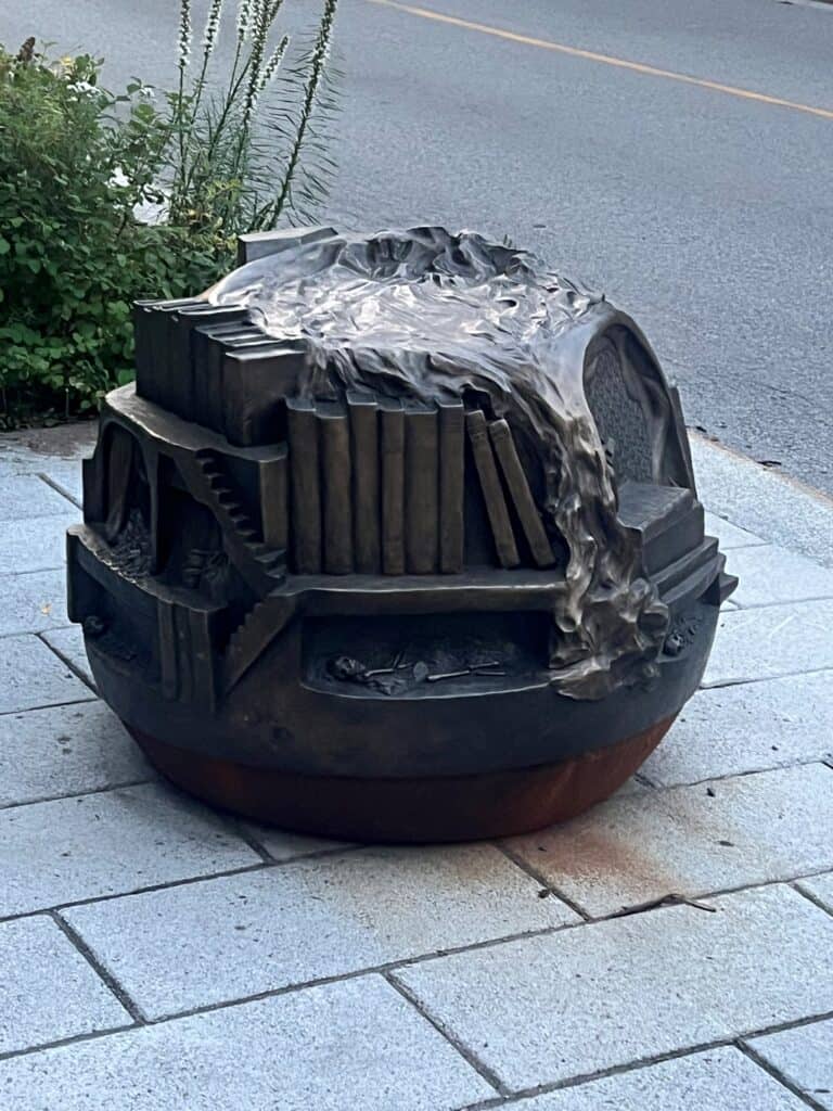 bronze sculpture bench on a sidewalk. The sculpture is roughly spherical, and includes books, tiny stairs, and other ephemera