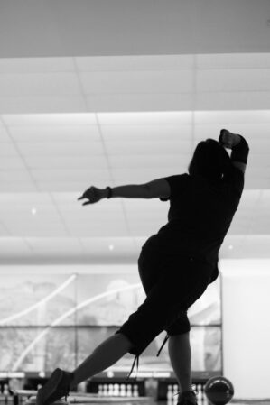 silhouette of a woman bowling. She is seen from behind, just after releasing the ball down the lane. The image is black and white.